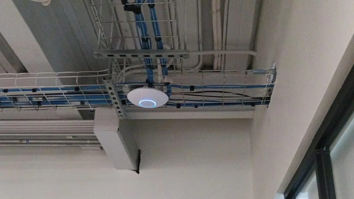 Punto de acceso WiFi montado bajo la escalerilla de cables de un pasillo, en una instalación de Tijuana
